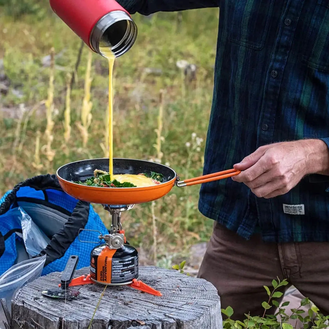 Lifestyle image of a man pouring beaten egg into a Jetboil Summit Skillet whilst being heated by a camping gas stove