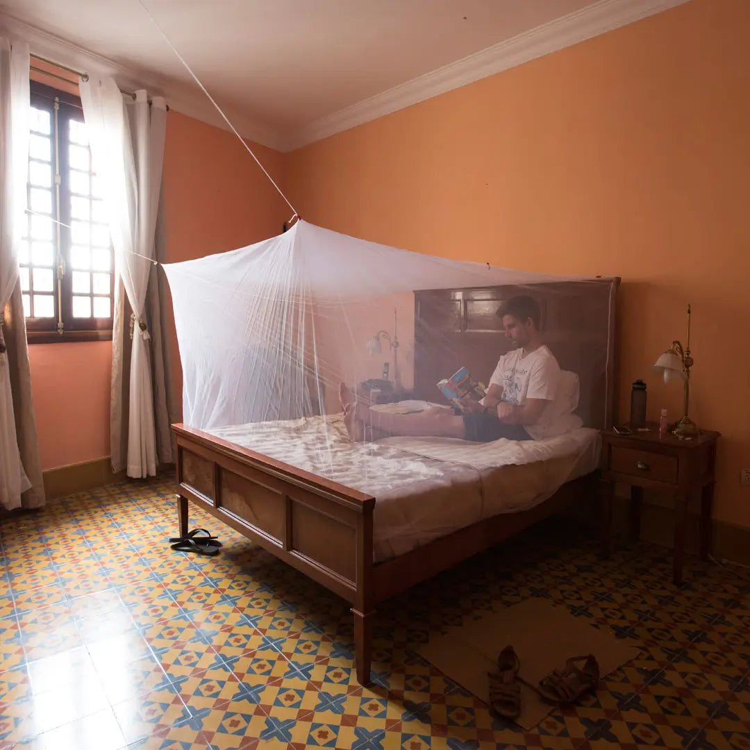 Person lying on a bed surrounded by a white Lifesystems boxnet mosquito net in a room with orange walls and tiled floor.