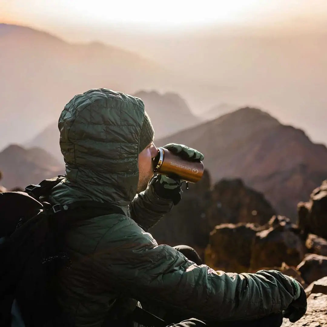 Person in a green jacket sitting on a mountain, drinking frm a Lifeventure Stainless Steel Thermal Mugin copper colour
