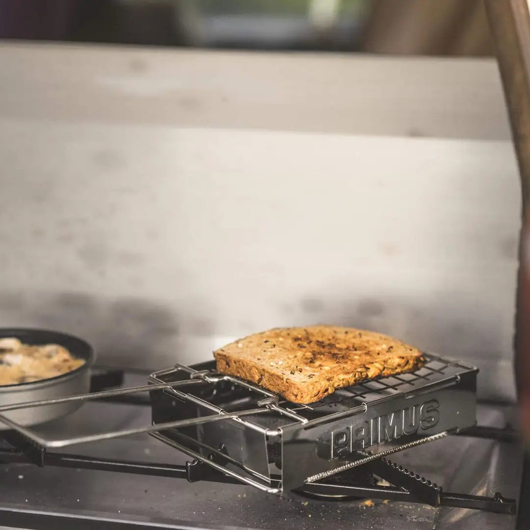 Primus toaster being used with a portable camping gas stove to make toast on a blurred background