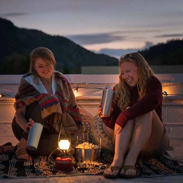 Two people sitting by a lit Primus Easy Light Duo gas lantern with drinks and snacks, enjoying a scenic view.