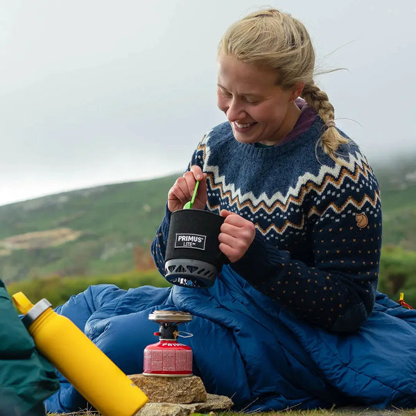 Outdoor image of a woman sittin ginside a sleeping bag whilst preparing a meal using a Primus Lite gas stove system