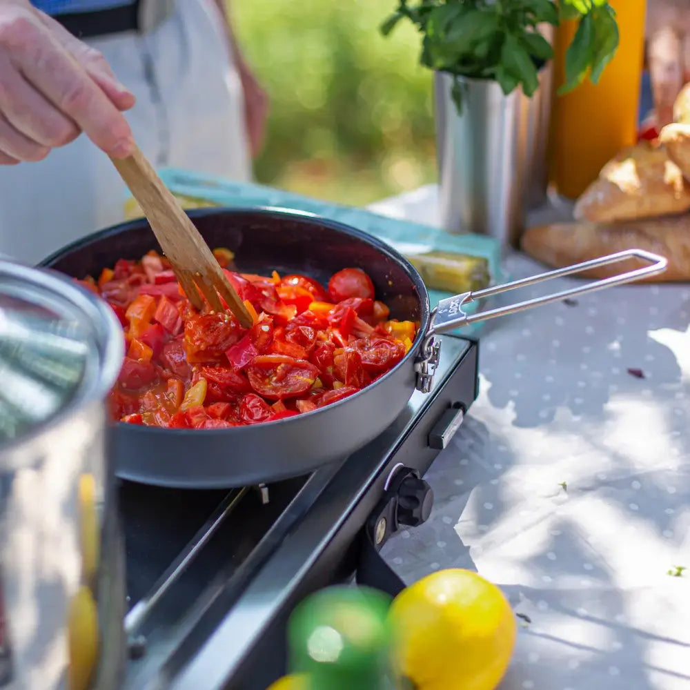 Person cooking tomatoes in a large Litech frying pan outside on a portable Primus camping stove 