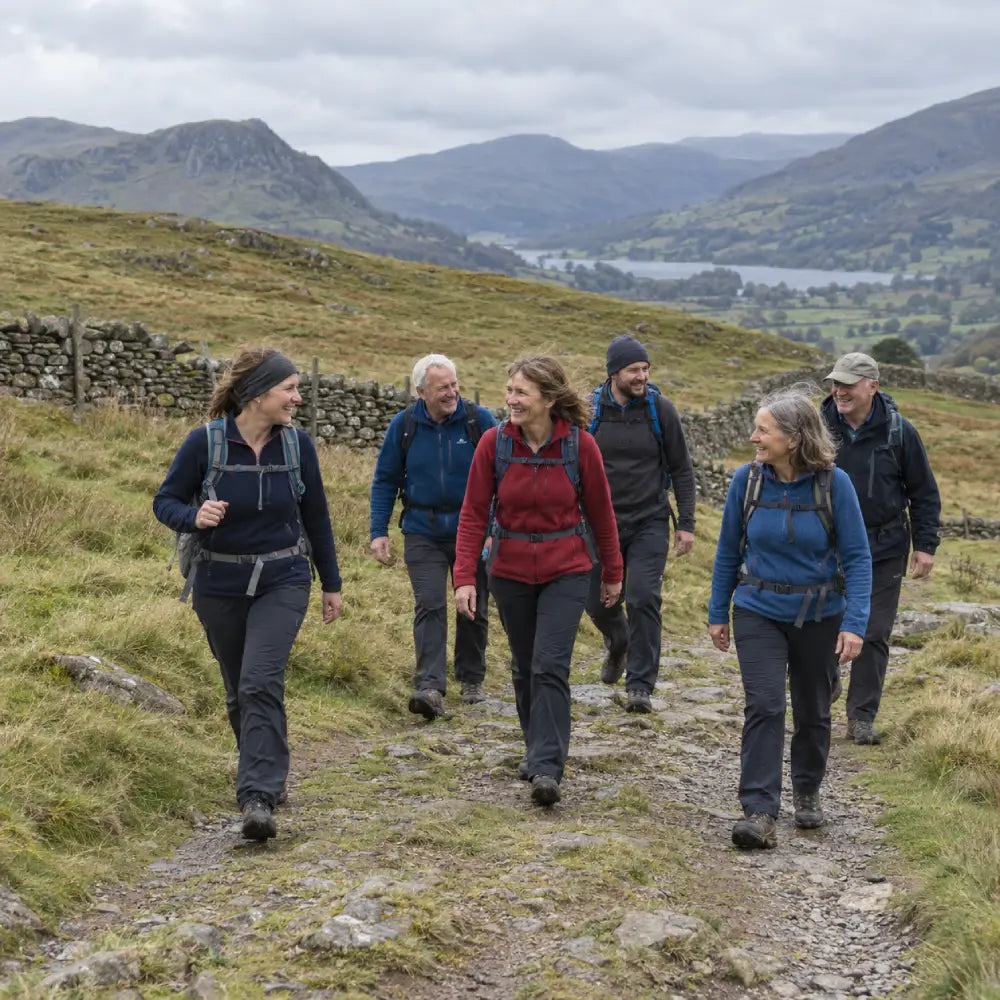 Six walkers with daypacks enjoying a countryside trail, with stone walls, hills and a lake in the background.