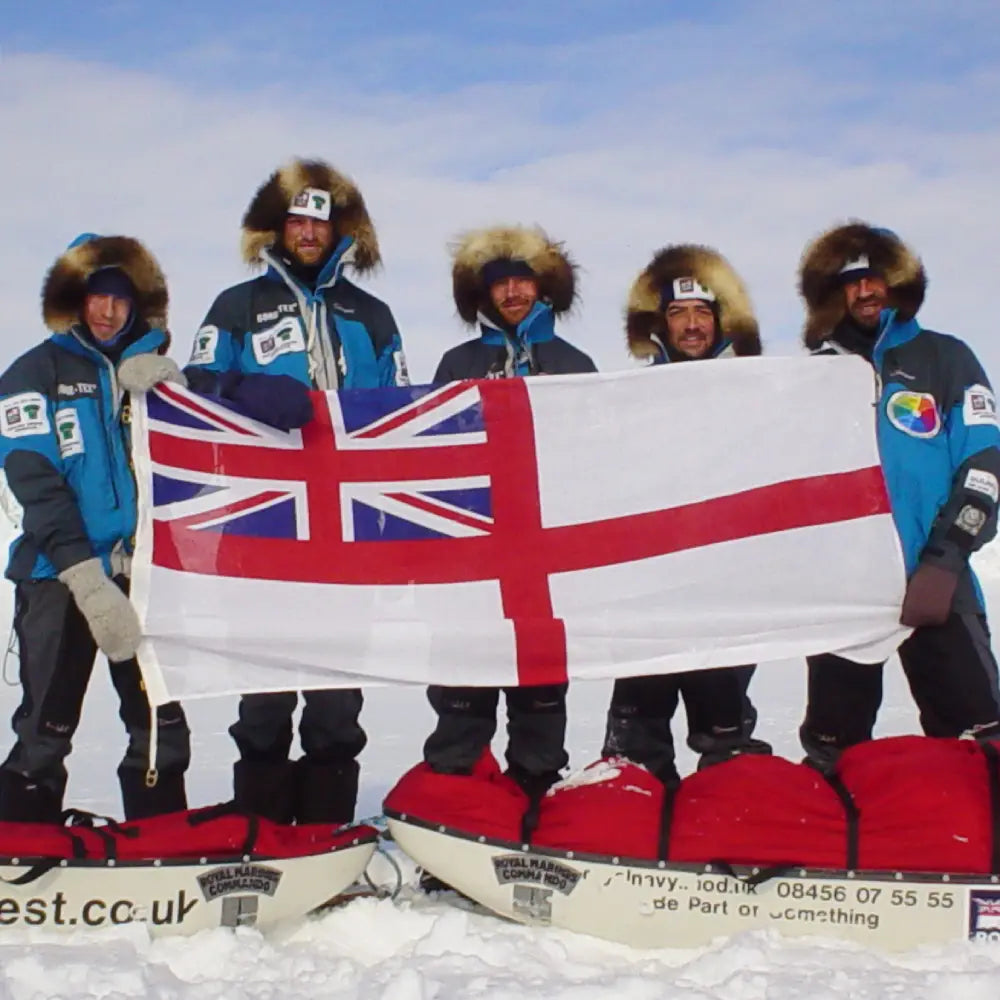 Five expedition team members in cold-weather clothing holding a White Ensign flag, with loaded sledges in the snow.
