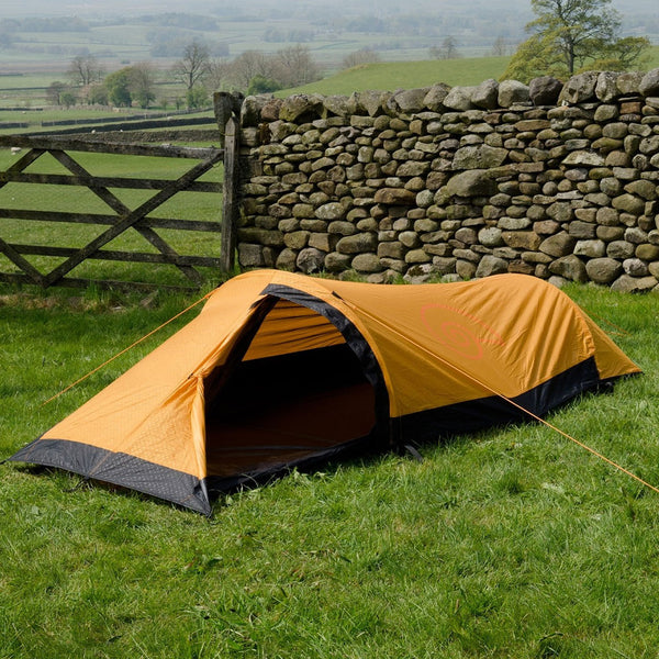 Lifestyle shot of of an orange Snugpak Journey solo bivi tent pitched in a field in front of a stone wall