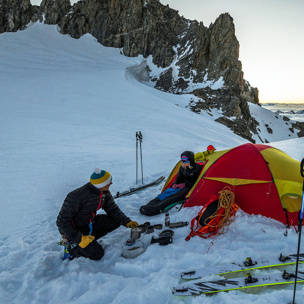 Lifestyle image of two alpine skiers setting up base camp  in the mountains surroundd by snow whilst wearing a Sub Zero Factor 2 plus thermal long sleeve mid layer top underneath their lightweight down jackets